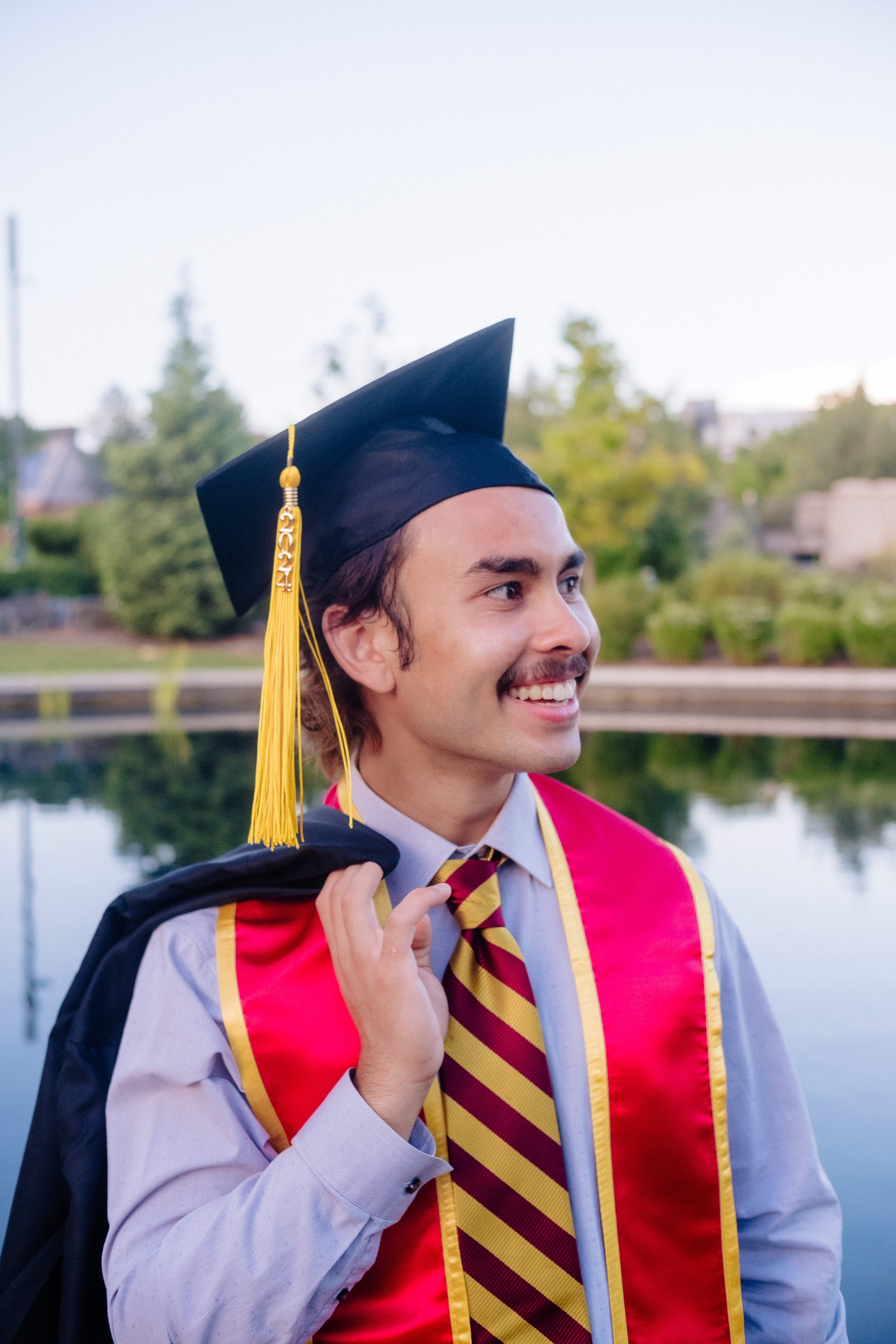 Graduate smiling in cap and gown with gold tassel by the campus lake
